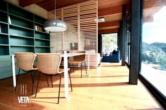 Interior de una casa moderna con mesa, sillas y grandes ventanales.